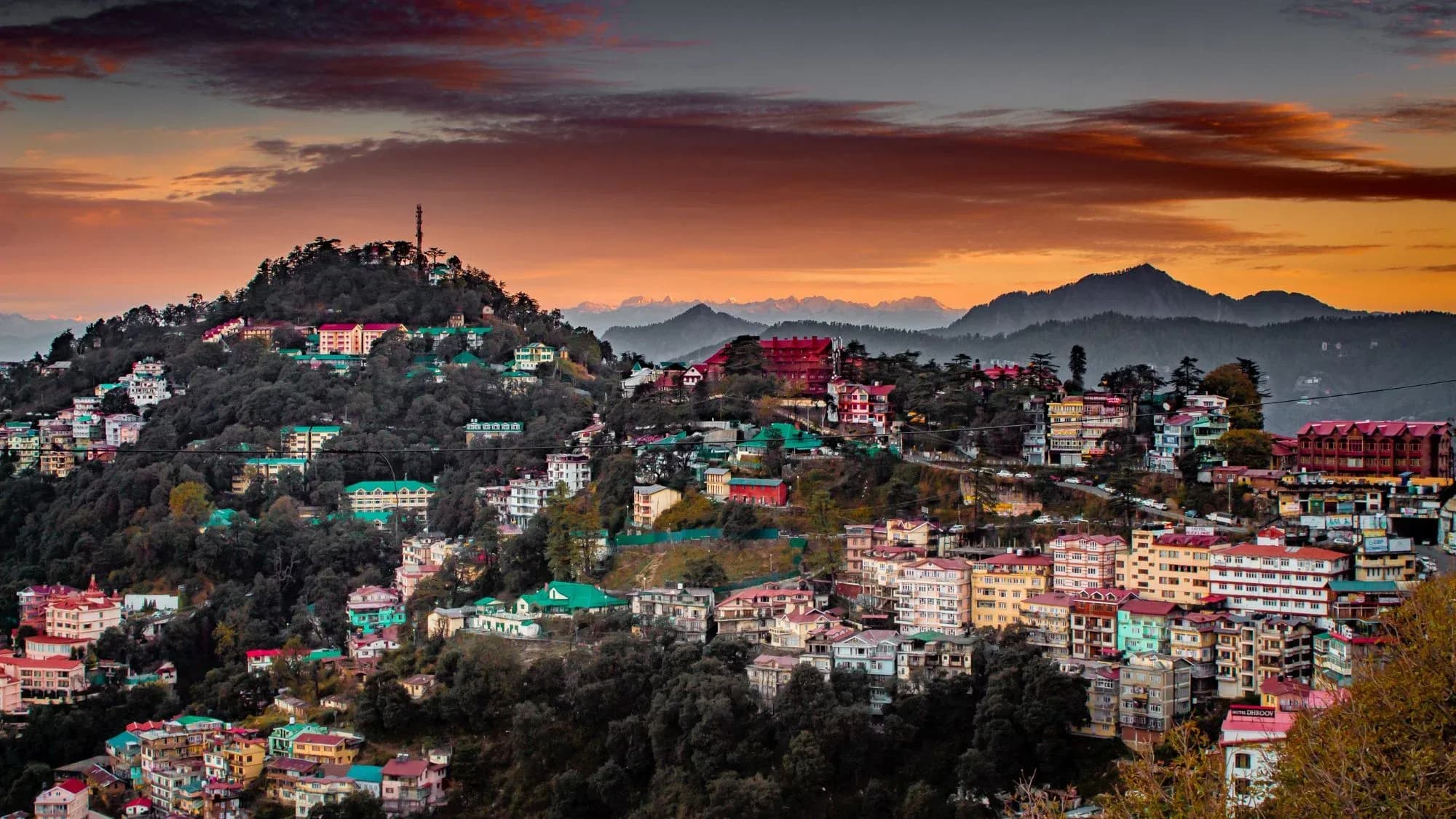 Colourful hillside houses of Shimla at sunset, with orange clouds and distant Himalayan peaks on the horizon