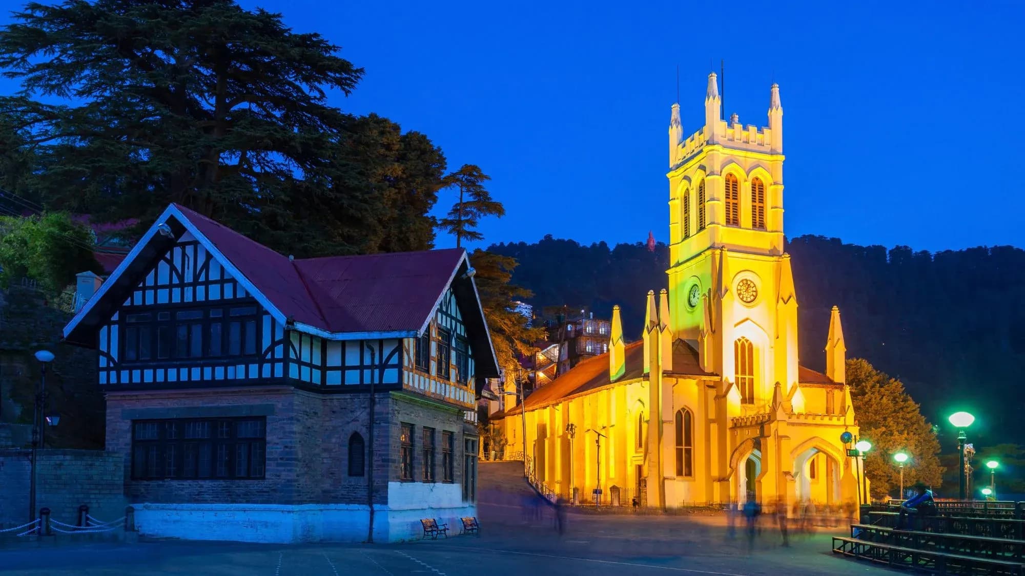 Christ Church in Shimla lit up in yellow at dusk, beside a black-and-white timbered building and cedar trees