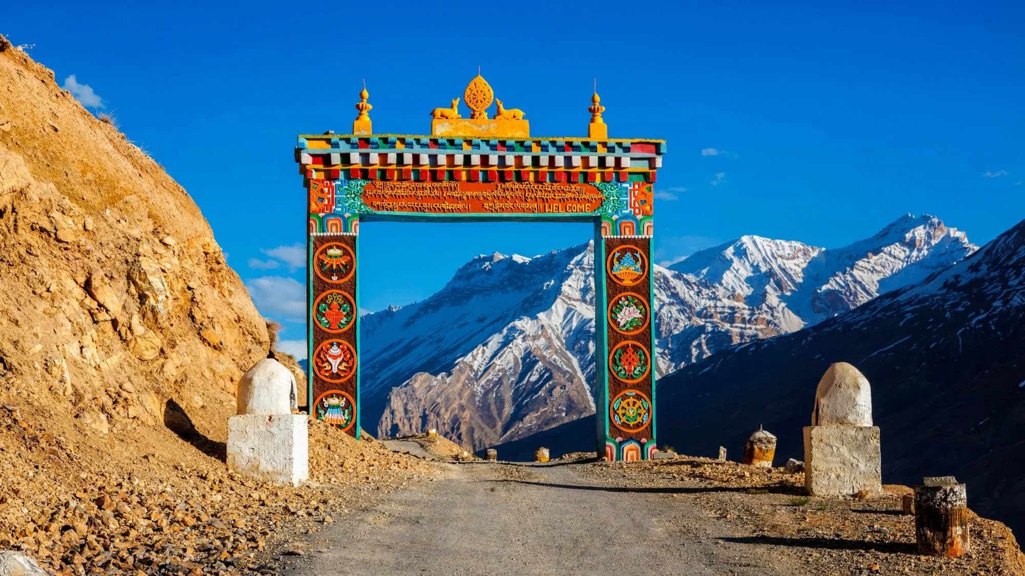 Colourful Tibetan-style welcome gate on a mountain road, framing snow-covered Himalayan peaks under a deep blue sky