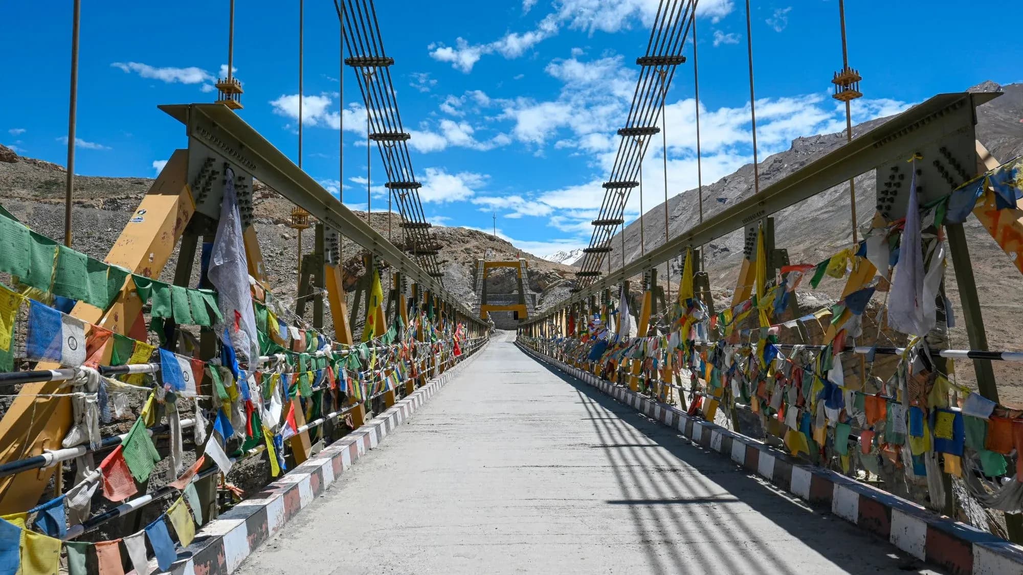 Suspension bridge hung with colourful prayer flags crossing a dry mountain valley under a bright blue sky
