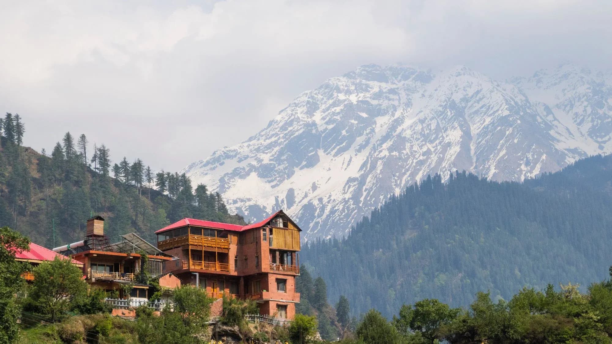 Wooden-balconied hill-stay building with a red roof below a snow-covered Himalayan peak and pine-forested slopes