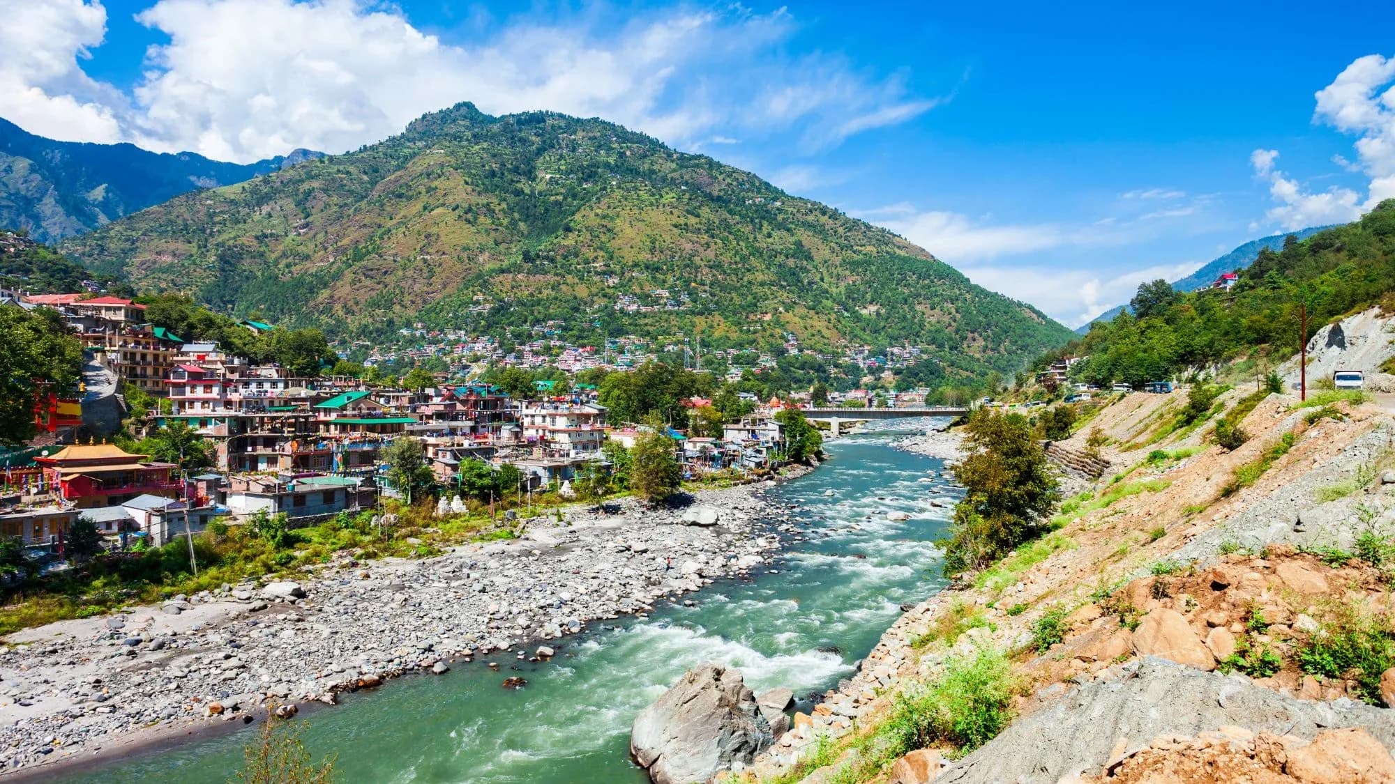 Himachal hill town on the banks of a turquoise mountain river, with a green forested mountain and blue sky behind