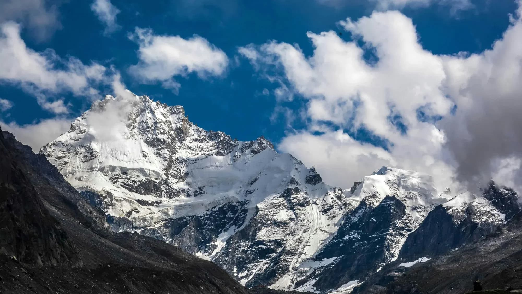 Glaciated snow-covered Himalayan peak rising above dark rocky slopes, with white clouds in a deep blue sky